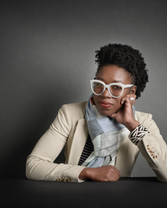 Dr. Joy Buolamwini sits at a table resting her head on her left hand. She wears white-framed glasses and a white blazer.