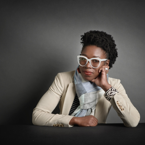 Dr. Joy Buolamwini sits at a table resting her head on her left hand. She wears white-framed glasses and a white blazer.